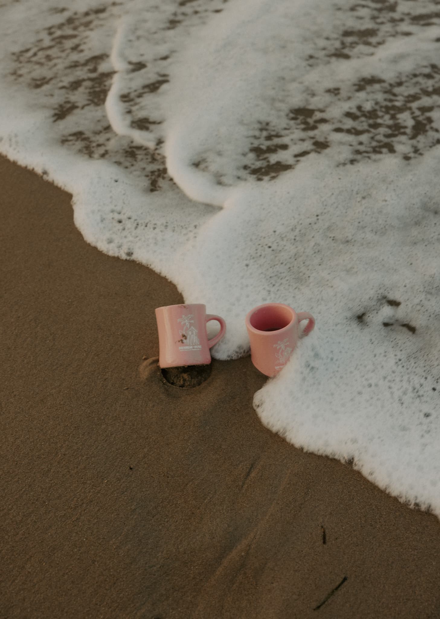 Two Daybreak Coffee Club diner mugs sitting in wet sand near the shore
