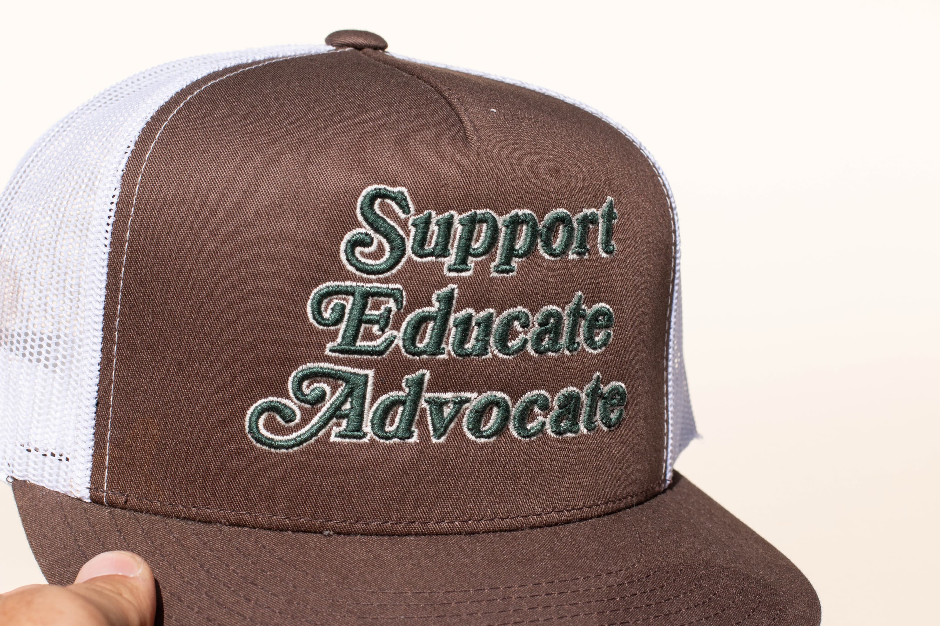 Brown and white trucker hat with raised Support Educate Advocate embroidery on the front panel